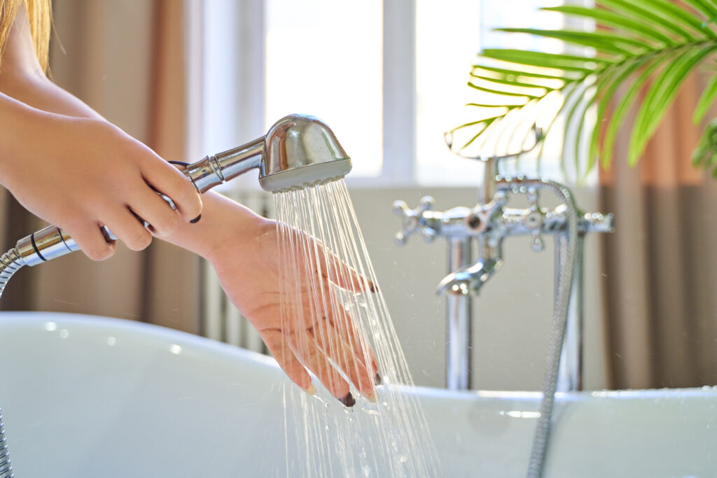 Water pours out of the shower on the girl’s hands. On a blue background. Woman checks the temperature of the water in the background of the bathroom